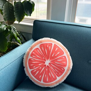 May include: A round, white pillow with a pink grapefruit slice design. The pillow is sitting on a teal blue couch.