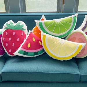 May include: A set of nine colorful fruit-shaped throw pillows, including a watermelon, strawberry, dragon fruit, raspberry, lime, lemon, fig, and grapefruit. The pillows are arranged on a blue couch.