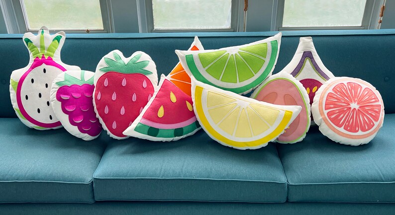 May include: A set of nine colorful fruit-shaped throw pillows, including a watermelon, strawberry, dragon fruit, raspberry, lime, lemon, fig, and grapefruit. The pillows are arranged on a blue couch.