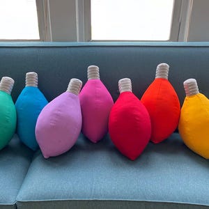 May include: Ten light bulb shaped pillows in various colors, including yellow, green, blue, purple, pink, red, orange, and white. The pillows are arranged in a row on a blue couch.