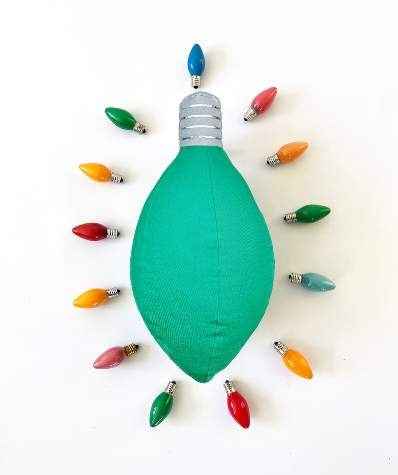 May include: A green lightbulb shaped pillow surrounded by 12 colorful lightbulbs. The lightbulbs are in various colors, including red, yellow, blue, and green.
