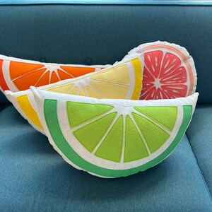 May include: Three decorative pillows shaped like citrus fruit slices. The pillows are stacked on top of each other. The top pillow is a pink grapefruit slice, the middle pillow is a yellow lemon slice, and the bottom pillow is a green lime slice.