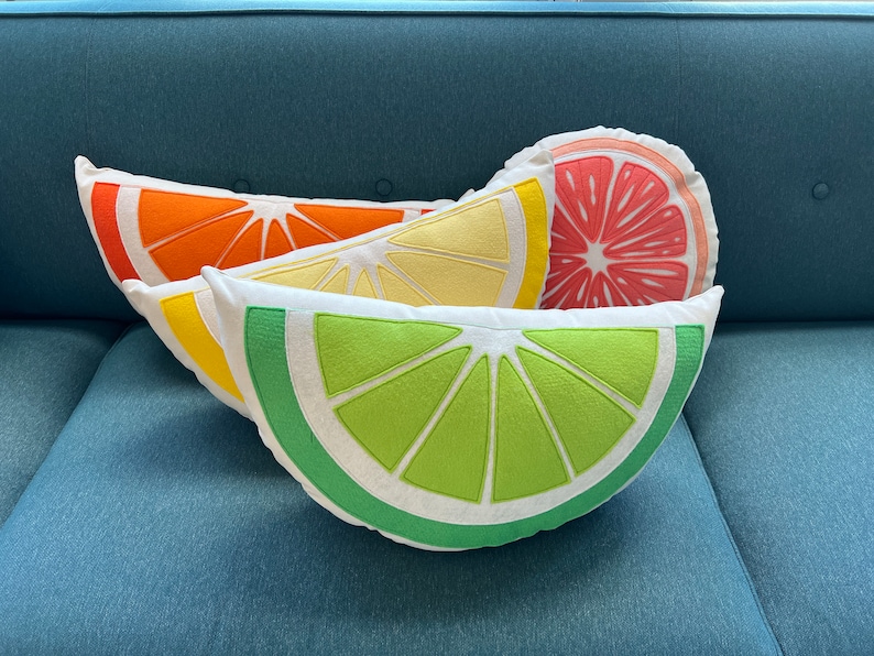 May include: Three decorative pillows shaped like citrus fruit slices. The pillows are stacked on top of each other. The top pillow is a pink grapefruit slice, the middle pillow is a yellow lemon slice, and the bottom pillow is a green lime slice.