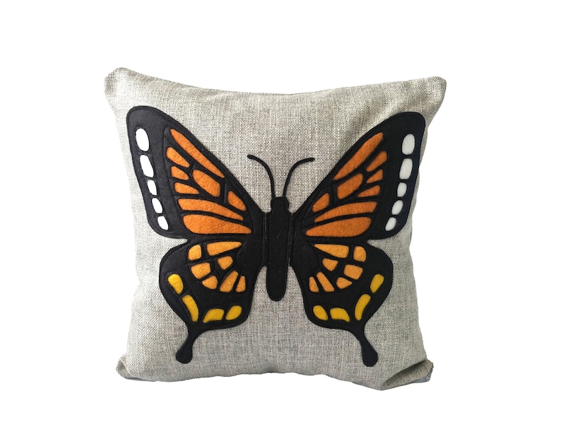 May include: A gray throw pillow with a large, black, orange, and yellow butterfly applique. The butterfly has a black body and wings with orange and yellow accents.