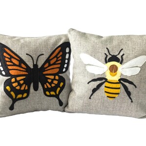 May include: Two decorative throw pillows with embroidered designs. One pillow features a black and orange butterfly with white accents. The other pillow features a yellow and black bee with white wings.