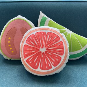 May include: Three decorative pillows shaped like fruit slices. The pillows are made of white fabric with embroidered details. The pillows are a pink grapefruit slice, a green lime slice, and a pink guava slice.