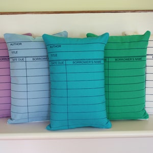 May include: Five decorative pillows with a library card design. The pillows are in various colors including purple, blue, teal, green, and white. The pillows have the words 'AUTHOR', 'TITLE', 'DATE DUE', 'BORROWER', and 'BORROWER'S NAME' printed on them.