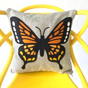 May include: A gray throw pillow with a large embroidered butterfly design. The butterfly is black, orange, and yellow.