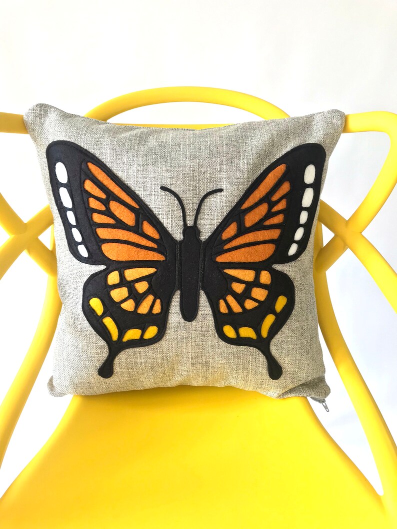 May include: A gray throw pillow with a large embroidered butterfly design. The butterfly is black, orange, and yellow.