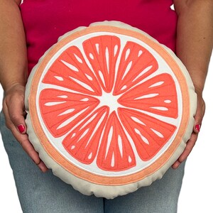 May include: A round, cream-colored pillow with a pink grapefruit slice design. The pillow is embroidered with white stitching.