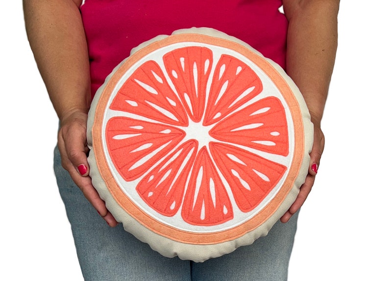 May include: A round, cream-colored pillow with a pink grapefruit slice design. The pillow is embroidered with white stitching.