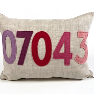 May include: A rectangular beige linen pillow with the numbers "07043" appliqued in shades of purple, red, and pink. The numbers are made of felt and are centered on the pillow.