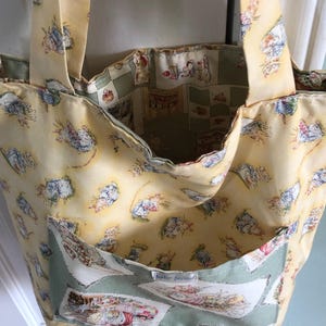 May include: A handmade tote bag with a yellow background and a repeating pattern of cartoon characters. The bag has two handles and a pocket with a light green border. The interior lining is a light green and white checkered pattern.