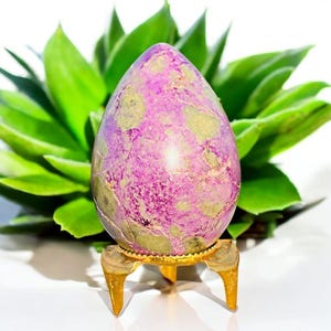 May include: A purple egg-shaped stone with green and gold markings, displayed on a small, gold-coloured stand. The background features bright green leaves. The stone is a decorative item.