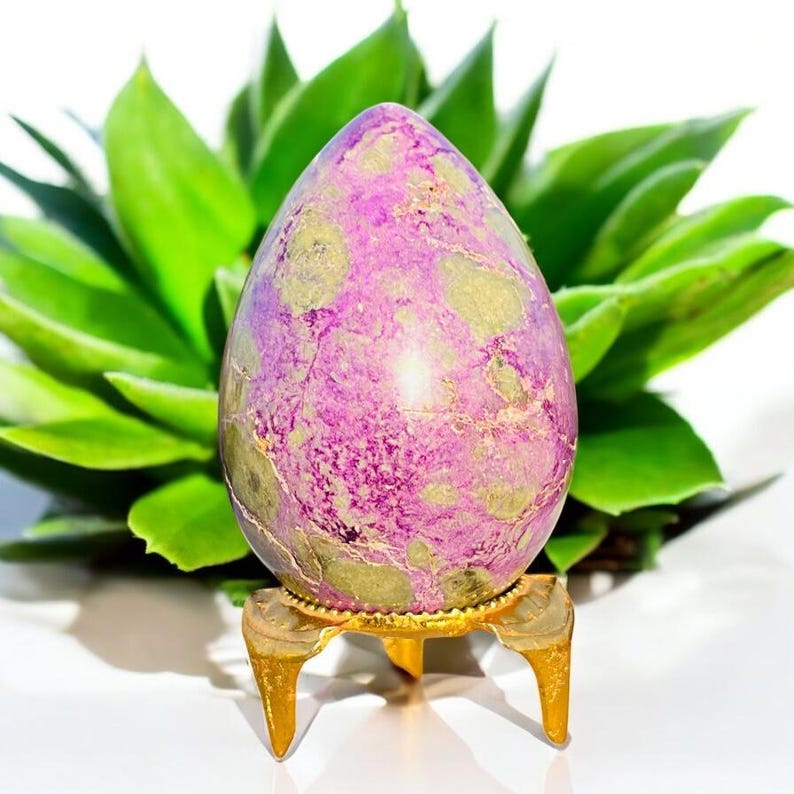 May include: A purple egg-shaped stone with green and gold markings, displayed on a small, gold-coloured stand. The background features bright green leaves. The stone is a decorative item.
