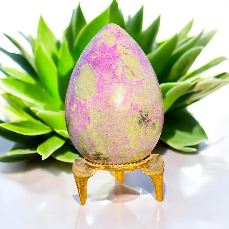 May include: An egg-shaped stone with a purple and green mottled pattern, presented on a small, gold-coloured stand with three legs. A green succulent plant is in the background. The stand is gold-coloured.