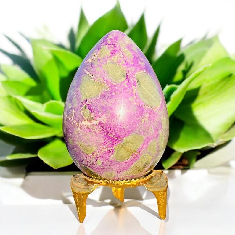 May include: A purple and green speckled egg-shaped stone, presented on a small gold stand with three legs. The stone is set against a backdrop of vibrant green leaves, creating a natural and elegant aesthetic.