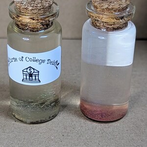 May include: Two small glass bottles with cork stoppers. The bottle on the left has a white label with the text "Curse of College Debt" and a small illustration of a building with a dollar sign. Both bottles contain a clear liquid with glitter.