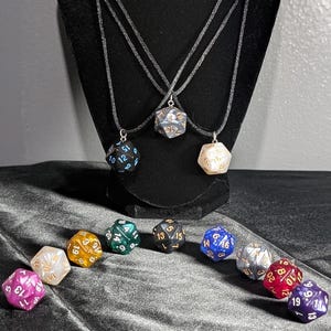 May include: A display of polyhedral dice necklaces and loose dice. The necklaces feature a variety of dice colors, including black, white, and blue, hanging on black cords. Loose dice in various colors are scattered below, including pink, green, and gold. The dice are used for tabletop games.