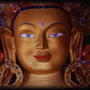 May include: Close-up of a golden Buddha statue with blue eyes and red lips. The statue has ornate headwear with floral and jewel accents. The background is dark, highlighting the statue's details.