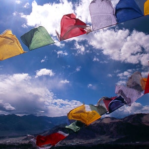 May include: A vibrant display of prayer flags in various colours, including red, yellow, and blue, set against a backdrop of a clear blue sky and fluffy white clouds. Mountains are visible in the distance.