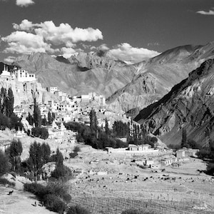 May include: Monochrome photograph of a mountain valley featuring a village and a monastery. The village is situated in the valley, with trees and buildings. Mountains are visible in the background.