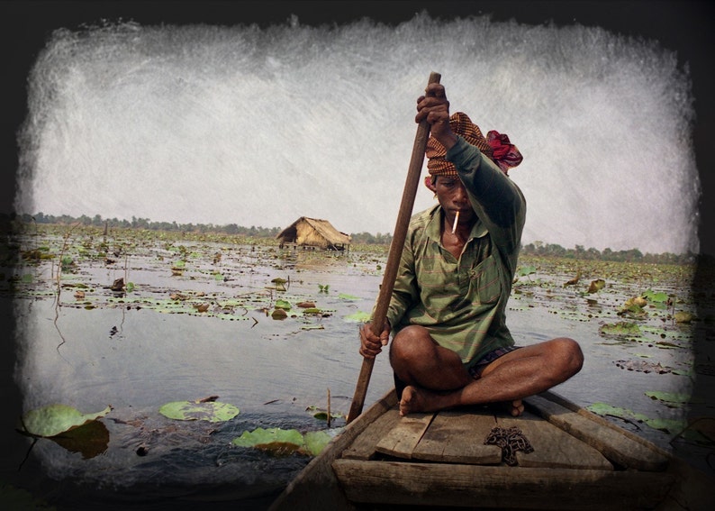 May include: A man rows a wooden boat through a lake filled with lily pads. He wears a green shirt and a patterned head covering. A small hut is visible in the distance. The image has a vintage, textured effect.