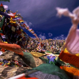May include: A scenic mountain view with vibrant prayer flags. The flags, in a variety of colours, are strung across the rocky landscape, set against a backdrop of a deep blue sky with scattered clouds.