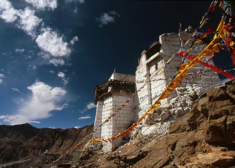 May include: A white stone building with a dark roof sits on a rocky hillside under a blue sky with clouds. Colourful prayer flags hang from the building. Mountains are visible in the background.