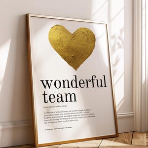 以下が含まれることがあります： 金色のハートと黒い文字で「wonderful team」と書かれた額入りのアートプリント。 プリントは金色のフレームで、白い壁と木の床に立てかけられています。