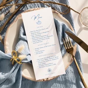 Può includere: Un menu con un design floreale blu è appoggiato su un piatto bianco, con una forchetta e un coltello dorati. Il menu elenca le opzioni di tè, le prelibatezze salate e i pasticcini e pasticceria. Sono presenti anche un portatovagliolo a fiocco dorato e un tovagliolo blu.