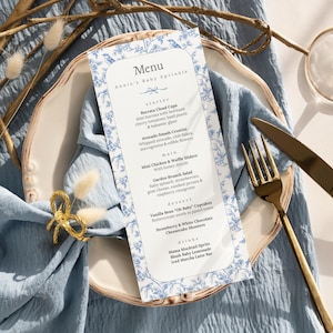 May include: A menu card with a blue floral border and text, "Annie's Baby Sprinkle," sits on a white plate atop a blue linen napkin. Gold cutlery and a wine glass are arranged nearby. Dried floral accents add to the elegant table setting.