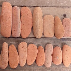 Può includere: Una collezione di quindici pietre lisce color terracotta disposte in due file orizzontali. Ogni pietra presenta una consistenza opaca e porosa con varie tonalità di arancione, marrone chiaro e rossastro, mostrando forme irregolari e allungate.