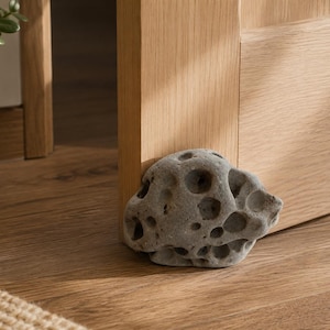 Pode incluir: Um batente de porta de pedra natural cinzenta e porosa, com uma forma arredondada irregular e numerosas pequenas e profundas reentrâncias na superfície, posicionado firmemente contra a base de uma porta de madeira clara para a manter aberta.