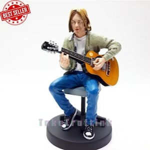 Op de afbeelding: Een beeldje van een muzikant die gitaar speelt, zittend op een kruk. De figuur draagt een blauwe spijkerbroek, een groene jas en zwart-witte sneakers. De gitaar is bruin en geel. De basis is zwart en een "Best Seller" sticker staat linksboven.