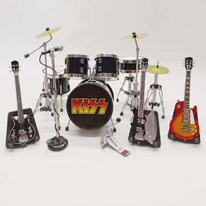 Op de afbeelding: Miniatuur KISS band set met een drumstel met het logo van de band, twee gitaren, een basgitaar, een microfoon en bekkens. De drumkit is zwart met zilveren hardware. De gitaren zijn zwart, rood en geel.