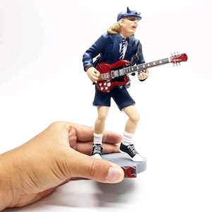 May include: A figurine of a musician playing a red electric guitar. The figure wears a blue school uniform, a cap with horns, and black and white trainers. The figurine is held by a hand.