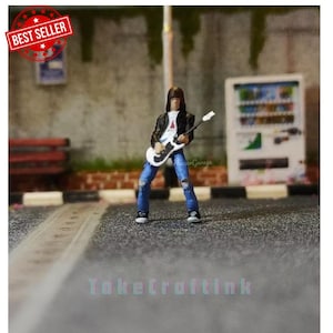May include: A miniature figurine of a person playing an electric guitar. The figurine is wearing a black leather jacket, blue jeans, and a white t-shirt. The guitar is white with black accents. The figurine is standing on a gray surface.