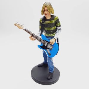 Op de afbeelding: Een beeldje van een persoon die een blauwe elektrische gitaar bespeelt. De figuur draagt een groen en zwart gestreept shirt, een blauwe spijkerbroek en zwarte schoenen. De gitaar heeft een zwarte slagplaat en een witte hals. Het beeldje staat op een zwarte cirkelvormige basis.