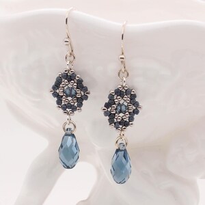 May include: Silver dangle earrings with a geometric design of dark blue and silver beads. The earrings feature a teardrop-shaped crystal in a dark blue color.