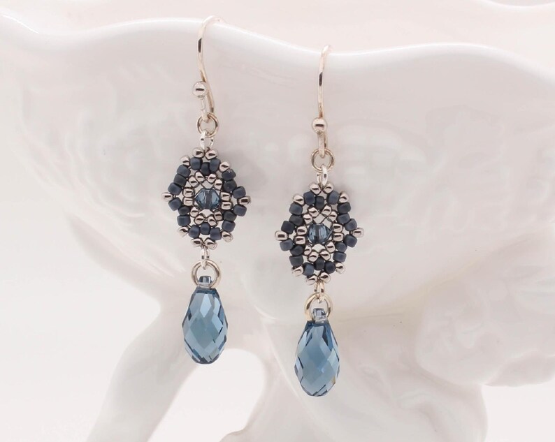 May include: Silver dangle earrings with a geometric design of dark blue and silver beads. The earrings feature a teardrop-shaped crystal in a dark blue color.