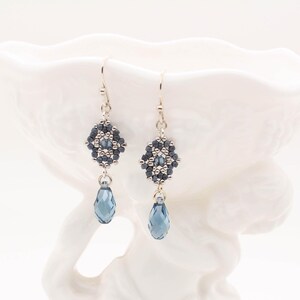 May include: A pair of silver dangle earrings with a geometric bead design in shades of gray and blue. The earrings feature a teardrop-shaped crystal in a dark blue color.