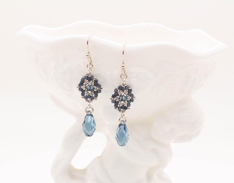 May include: A pair of silver dangle earrings with a geometric bead design in shades of gray and blue. The earrings feature a teardrop-shaped crystal in a dark blue color.