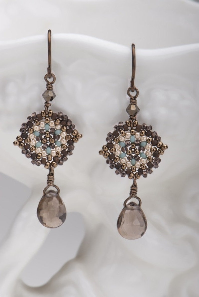 May include: A pair of dangle earrings with a geometric design made of brown, white, and blue beads. The earrings have a teardrop-shaped brown gemstone hanging from the bottom.