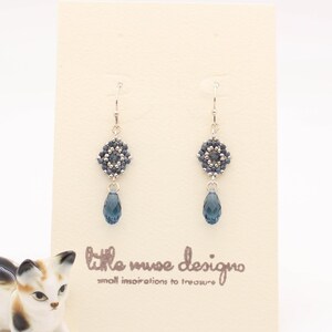 May include: A pair of silver dangle earrings with a blue crystal bead and a beaded flower design. The earrings are displayed on a white card with the text "little muse design" and "small inspirations to treasure".