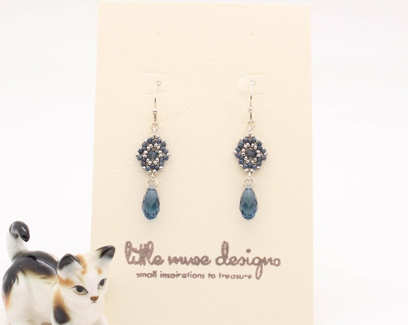 May include: A pair of silver dangle earrings with a blue crystal bead and a beaded flower design. The earrings are displayed on a white card with the text "little muse design" and "small inspirations to treasure".