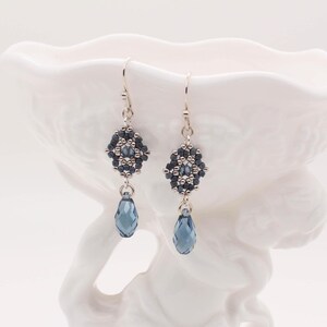 May include: A pair of silver dangle earrings with a geometric design made of dark blue and silver beads. Each earring features a teardrop-shaped blue crystal.