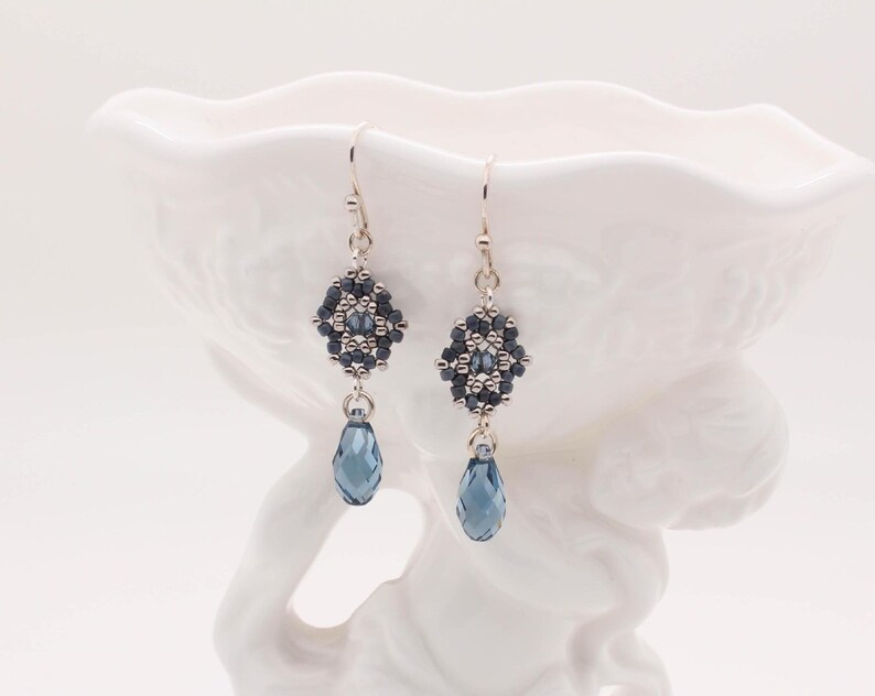 May include: A pair of silver dangle earrings with a geometric design made of dark blue and silver beads. Each earring features a teardrop-shaped blue crystal.