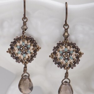 May include: A pair of dangle earrings with a geometric design made of brown, white, and blue beads. The earrings have a teardrop-shaped smoky quartz gemstone hanging from the bottom.