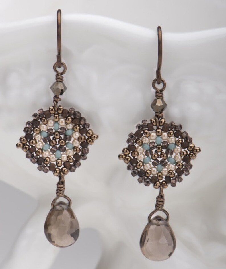 May include: A pair of dangle earrings with a geometric design made of brown, white, and blue beads. The earrings have a teardrop-shaped smoky quartz gemstone hanging from the bottom.